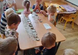 Dzieci zgromadzone wokół stołu obserwują, jak nauczycielka wkłada mus do słoika